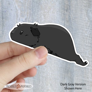 May include: A dark gray cartoon guinea pig sticker. The guinea pig is lying on its side with its head turned to the left. The sticker is being held by a hand.