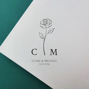 May include: A simple black line drawing of a rose with leaves, the initials "C" and "M", the names "Clare & Michael", and the date "12.07.2026" printed in black on a white background. The design is on a white card.