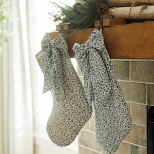 Puede incluir: Dos calcetines de Navidad blancos y azules con estampado floral y lazos colgados de una repisa de madera sobre una chimenea de azulejos.