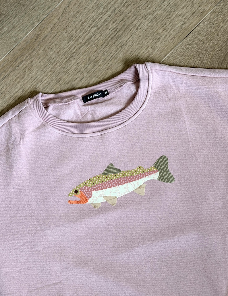 May include: A light purple sweatshirt with a crew neck and a fish applique. The fish has a white belly, pink and green patterned body, and orange and yellow accents. The brand name "EasyGala" is on the tag.