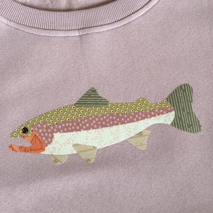 May include: A mauve sweatshirt featuring an appliqued fish design. The fish is composed of various patterned fabrics in shades of green, pink, and beige. The fish has an orange mouth and a striped tail. The sweatshirt has a ribbed neckline.