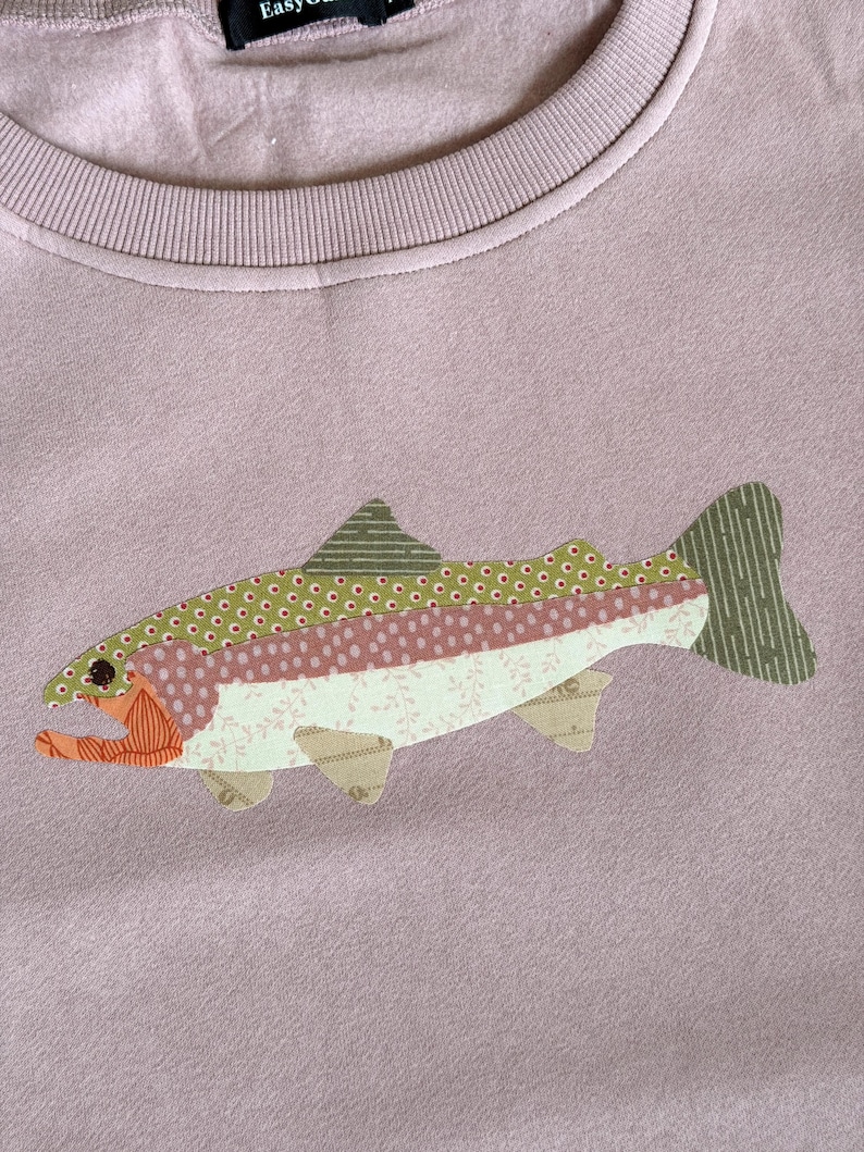 May include: A mauve sweatshirt featuring an appliqued fish design. The fish is composed of various patterned fabrics in shades of green, pink, and beige. The fish has an orange mouth and a striped tail. The sweatshirt has a ribbed neckline.