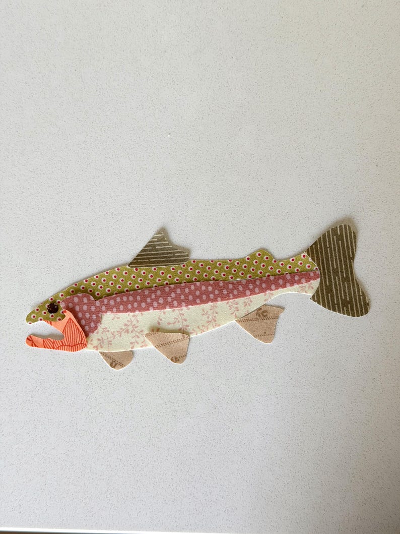 May include: A colorful fish applique made from patterned fabric. The fish features a green polka dot top, a pink floral middle, and a cream floral bottom. The fins and tail are brown with vertical stripes.