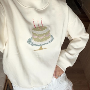 May include: A cream-colored long-sleeve sweatshirt featuring a mock neck collar and a central embroidered graphic of a multi-layered cake on a pedestal stand. The cake design includes three lit candles and various patterned fabric textures.
