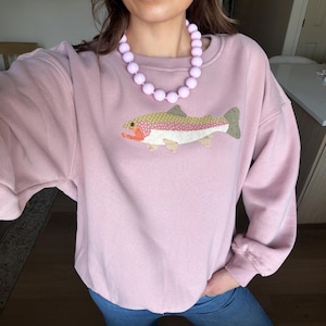 May include: A light pink sweatshirt with a colorful embroidered fish design. The sweatshirt is paired with blue jeans and a light purple beaded necklace. The fish has a yellow, pink, and green pattern with orange and white accents.