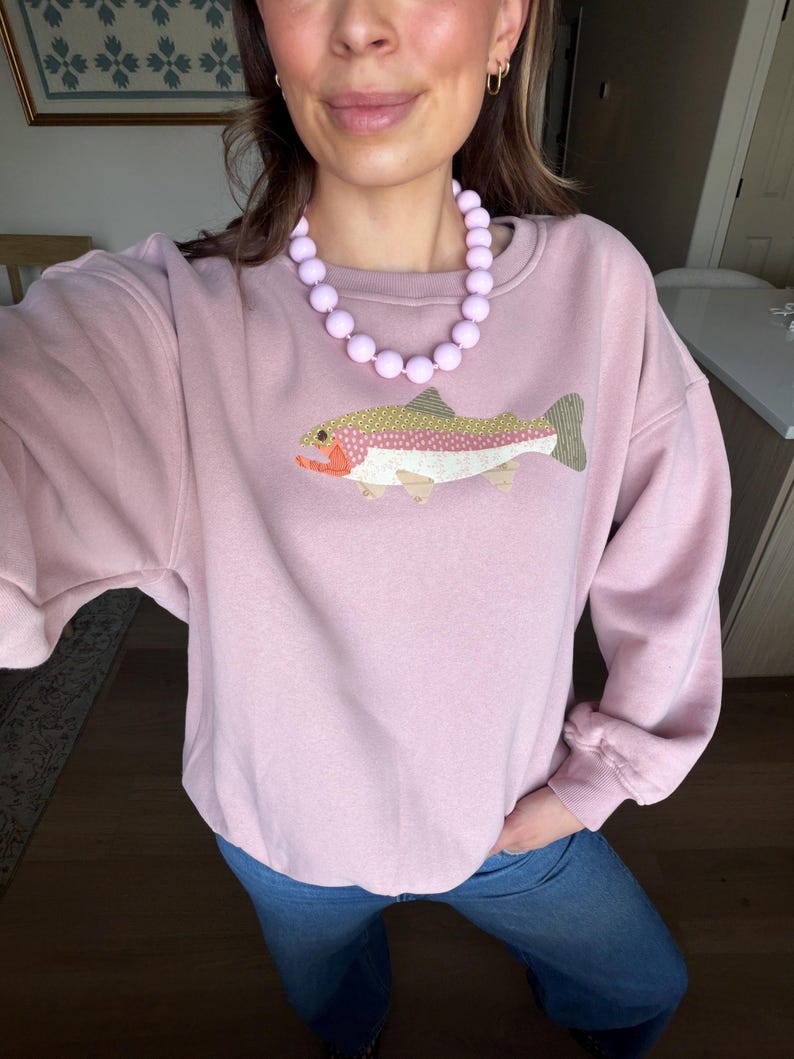May include: A light pink sweatshirt with a colorful embroidered fish design. The sweatshirt is paired with blue jeans and a light purple beaded necklace. The fish has a yellow, pink, and green pattern with orange and white accents.