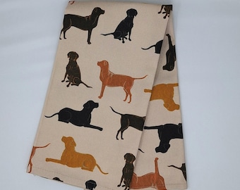 May include: A folded beige tea towel with a repeating pattern of dog silhouettes in black, brown, and orange. The dogs are shown in different poses, including standing, sitting, and lying down. The tea towel is made of a soft fabric.