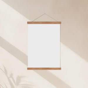 May include: A blank white poster hanging from wooden bars, suspended by a thin string. The poster is rectangular and centered, with the wooden bars at the top and bottom. The background is a light beige wall with shadows.