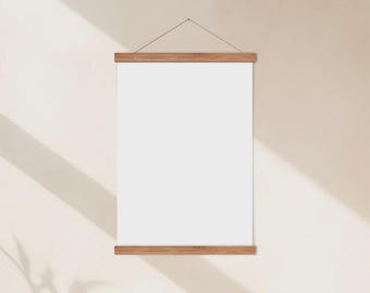 May include: A blank white poster hanging from wooden bars, suspended by a thin string. The poster is rectangular and centered, with the wooden bars at the top and bottom. The background is a light beige wall with shadows.