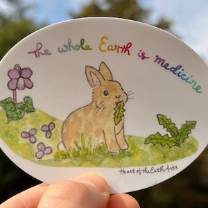 May include: A white oval sticker with a watercolor illustration of a brown rabbit eating a green leaf. The text "The whole Earth is medicine" is written in rainbow colors above the rabbit. The text "Heart of the Earth Arts" is written in black at the bottom of the sticker.