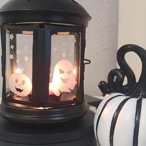 May include: Black Halloween lantern with pumpkin and ghost cutouts, lit by a candle. A white pumpkin with black stripes sits next to the lantern. The lantern has a round base and a handle on top.