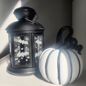 May include: A black metal lantern with bat and star cutouts next to a white pumpkin with black vertical stripes and a black curly stem. The lantern has a handle and a rounded top. The pumpkin is a decorative item.