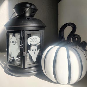 May include: A black metal lantern with ghost decals and the word "BOO!" in a speech bubble, next to a white pumpkin with black stripes and a black stem. The lantern has a handle and star cutouts.
