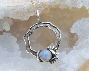 Puede incluir: Un collar de plata con un colgante de piedra lunar. El colgante es un círculo con un diseño de sol en un lado y una piedra lunar en el centro.