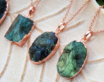 May include: Four necklaces with raw, iridescent labradorite pendants set in rose gold-colored metal. The pendants vary in shape and are suspended from delicate chains. The necklaces are displayed on a white, leaf-shaped dish.