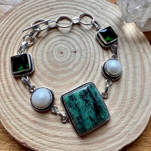 Pode incluir: Uma pulseira de prata com pedras preciosas quadradas e redondas. A pulseira tem uma grande pedra verde quadrada no centro, rodeada por pedras verdes e brancas menores. A pulseira é exibida em uma superfície de madeira.
