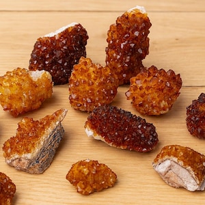 Op de afbeelding: Een verzameling ruwe, ongeslepen citrienkristallen in tinten amber, bruin en goud. De kristallen hebben een ruw, getextureerd oppervlak en zijn gerangschikt op een licht houten oppervlak. De kristallen variëren in grootte en vorm.