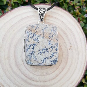 May include: A silver pendant with a rectangular stone featuring a dendritic agate pattern in shades of brown, white, and blue. The pendant is on a black cord.
