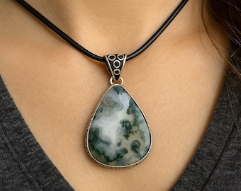 May include: A teardrop-shaped pendant necklace featuring a natural stone with green and white patterns. The stone is set in a silver-colored metal frame and hangs from a black cord necklace.
