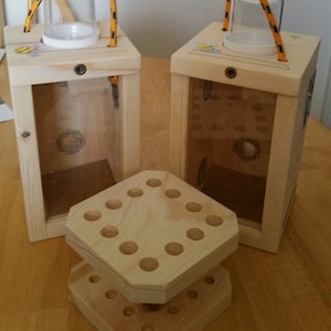 May include: Two wooden beekeeping boxes with clear plastic windows and a rope handle. The boxes are empty and have a small hole in the side. A wooden stand with holes is in front of the boxes.