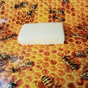May include: A white rectangular bar of soap on a yellow and orange honeycomb background with bees.