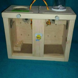 May include: Wooden toy box with two compartments, each with a small opening and a mesh screen. The box has a rope handle and a bee sticker on the front. There is a small clear plastic container on top of the box.