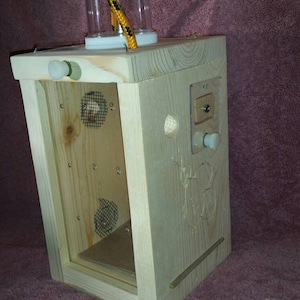 May include: A wooden box with a clear plastic container on top, a mesh screen on the inside, and a knob on the side. The box is made of light wood and has a natural finish.