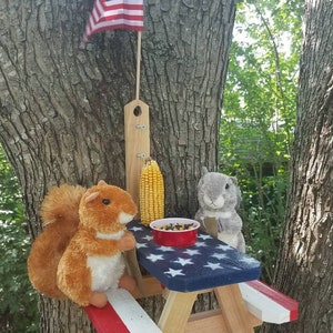 May include: A red, white, and blue picnic table bird feeder with a squirrel and a chipmunk sitting on it. The table is attached to a tree and has a small American flag hanging above it.