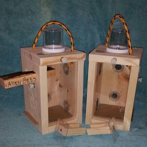 May include: Two wooden boxes with clear plastic containers and rope handles. The boxes have a mesh screen on one side and a small wooden door on the other. One box has the words "Alien Bees" written on it.