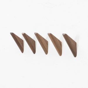 May include: Five wooden wall hooks in a row, each with a triangular shape and a natural brown finish. The hooks are mounted on a white wall, showcasing their minimalist design. These hooks are ideal for hanging coats, hats, or other items.
