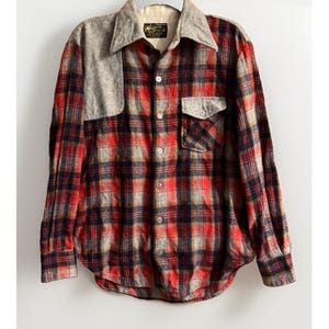 May include: A long-sleeve button-down shirt featuring a red, black, and gray plaid pattern. The shirt includes a solid gray shoulder yoke and a matching gray chest pocket. A white label with black text is visible on the inside collar area.