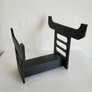 May include: Black plastic stand with a shelf and a raised back. The stand is designed to hold a gaming console.