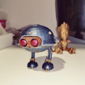 Pode incluir: Um brinquedo de robô azul e dourado com lentes oculares vermelhas e uma pequena estatueta dourada de um personagem. O robô tem uma cabeça arredondada, duas pernas e dois pés. A estatueta está sentada com os braços estendidos.