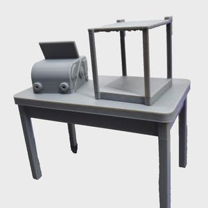 Puede incluir: Una mesa gris en miniatura con una caja registradora y un expositor. La mesa tiene cuatro patas y una parte superior rectangular. La caja registradora tiene una pantalla y dos perillas. El expositor es un pequeño marco cuadrado.