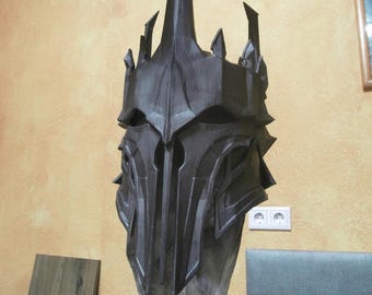 May include: A dark gray, stylized helmet with a tall central spike and multiple smaller spikes. The helmet has a detailed, angular design and is mounted on a stand. The background is a light yellow wall.