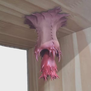 Puede incluir: Una escultura biomórfica surrealista, en tonos rosa y rojo, que emerge de una superficie de madera. La obra de arte presenta una forma orgánica texturizada con protuberancias afiladas y puntiagudas, creando un contraste visual llamativo. La pieza evoca lo extraño y lo de otro mundo.