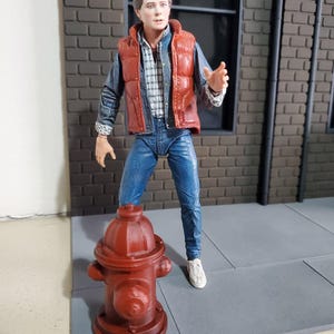Op de afbeelding: Een beweegbaar actiefiguur van een jong persoon met bruin haar, gekleed in een rood gewatteerd vest over een geruit overhemd, spijkerjas en blauwe spijkerbroek, staand naast een klein, donkerrood model van een brandkraan.