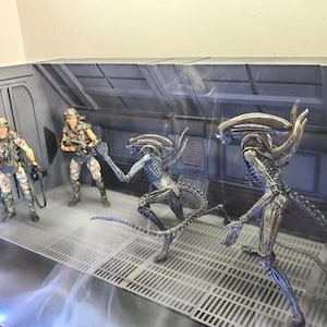 Könnte beinhalten: Ein Diorama mit Actionfiguren aus dem Film Alien. Zwei Figuren in Militäruniformen stehen vor zwei Xenomorph-Figuren. Die Szene spielt in einer metallischen, industriellen Umgebung.