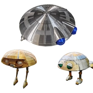 Pode incluir: Três modelos impressos em 3D de naves espaciais. O maior é prateado com detalhes azuis. Dois modelos menores são dourados e bege, cada um com um design único e pequenas pernas.