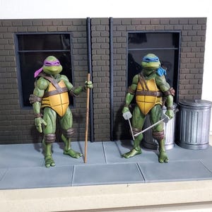 May include: Two Teenage Mutant Ninja Turtles action figures standing on a gray sidewalk diorama. One figure wears a purple mask and holds a wooden staff, while the other wears a blue mask and holds two silver swords. A red fire hydrant and trash can are included.