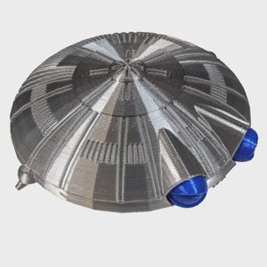 May include: A 3D-printed, silver-coloured, disc-shaped object with a detailed geometric pattern. The object has two blue, spherical protrusions on the bottom. The design suggests a futuristic or sci-fi theme.