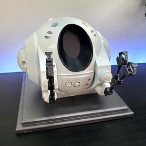 May include: A white, spherical, 3D-printed model with a large, dark opening and two articulated black arms. The model has several small, circular indentations and is mounted on a gray rectangular base. The design suggests a futuristic or robotic theme.