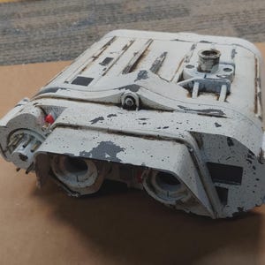 May include: A weathered, off-white rectangular device with visible wear and tear. The object has a complex design with multiple openings, protrusions, and a small red button. The surface shows signs of paint chipping, revealing darker layers beneath.