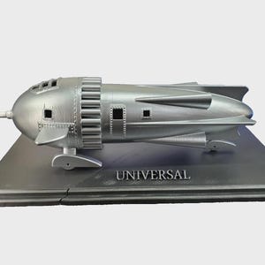 May include: A silver model of a retro-futuristic rocket ship, mounted on a rectangular base. The rocket has a streamlined design with fins, windows, and a pointed nose. The word "UNIVERSAL" is printed on the base.