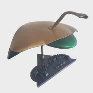 May include: A gold and green sculpture of a flying saucer with a curved antenna and a black base. The base has the text "War of the Worlds 1953" in a stylized font. The sculpture is set against a white backdrop.