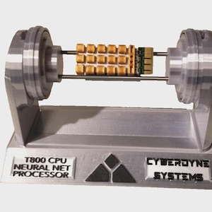 Puede incluir: Modelo plateado de un procesador de red neuronal T800 CPU. El modelo presenta una unidad central de procesamiento con componentes dorados y el texto "CYBERDYNE SYSTEMS". La base tiene el texto "T800 CPU NEURAL NET PROCESSOR".