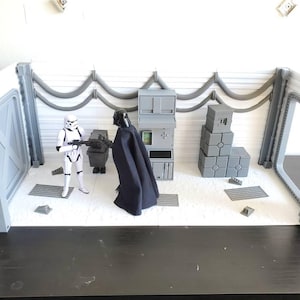Könnte beinhalten: Ein Star Wars Diorama mit Stormtrooper- und Darth Vader-Figuren. Die Szene umfasst graue Wände, eine Tür und verschiedene graue Strukturen, die eine detaillierte Miniaturumgebung schaffen. Die Figuren befinden sich in einem weißen Raum.