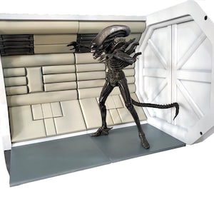 Op de afbeelding: Een gedetailleerde, donkergekleurde Alien-figuur staat in een futuristische, wit en grijs interieursetting. De figuur heeft een lange, gesegmenteerde staart en poseert met uitgestrekte armen. De achtergrond omvat een deur en wandpanelen.
