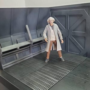 Puede incluir: Un detallado conjunto de diorama con una figura con un abrigo largo gris, pantalones marrones y zapatillas deportivas. El conjunto tiene un interior gris oscuro con bancos y una gran puerta. El suelo es una superficie negra cuadriculada.