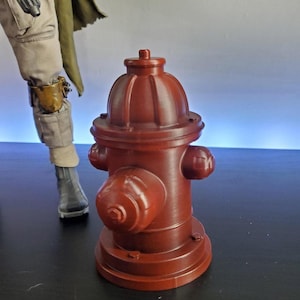 Op de afbeelding: Een donkerrood brandkraanmodel met een cilindrisch lichaam, een koepelvormige bovenkant en drie uitstekende dopjes. De brandkraan heeft een ronde voet met zichtbare boutdetails en een gladde, matte afwerking, rechtopstaand als object.